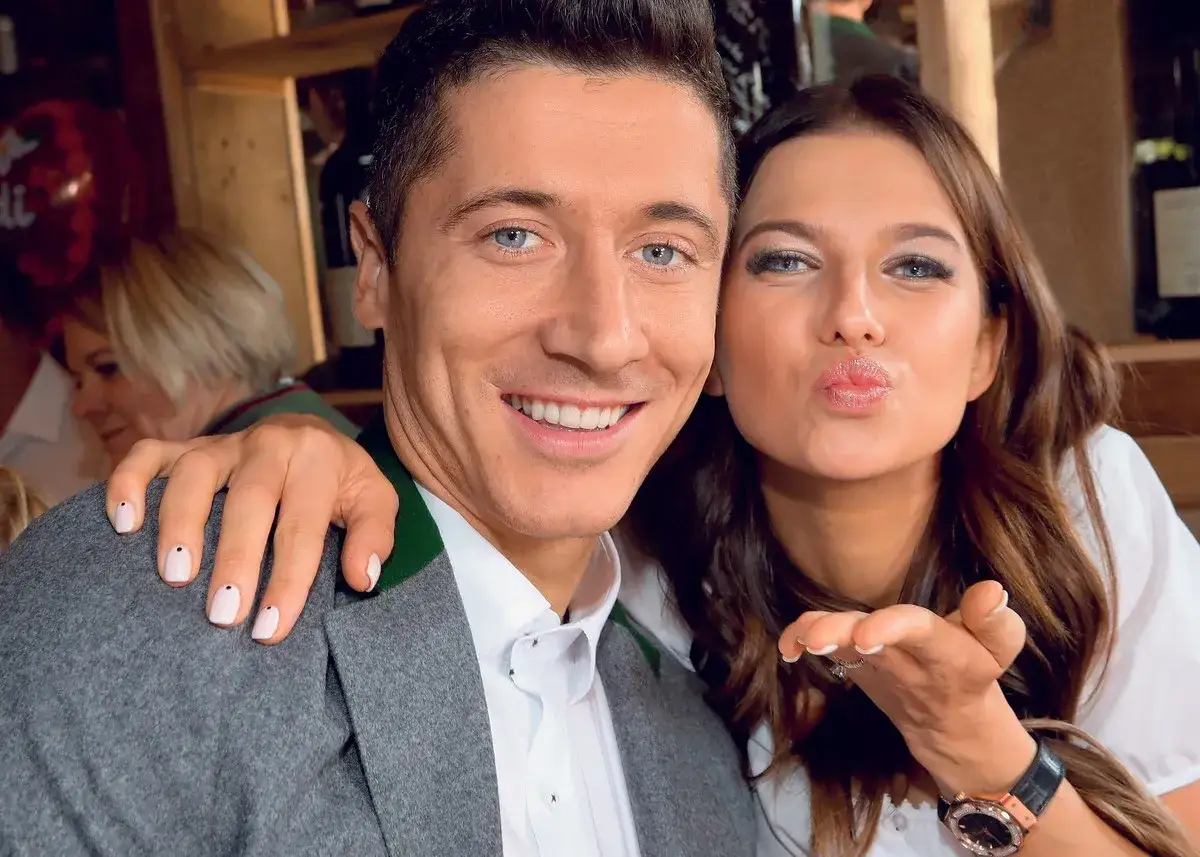 Uśmiechnięci Lewandowscy pozdrawiają z imprezy. Anna Lewandowska całuje, Robert Lewandowsk i uśmiecha się.