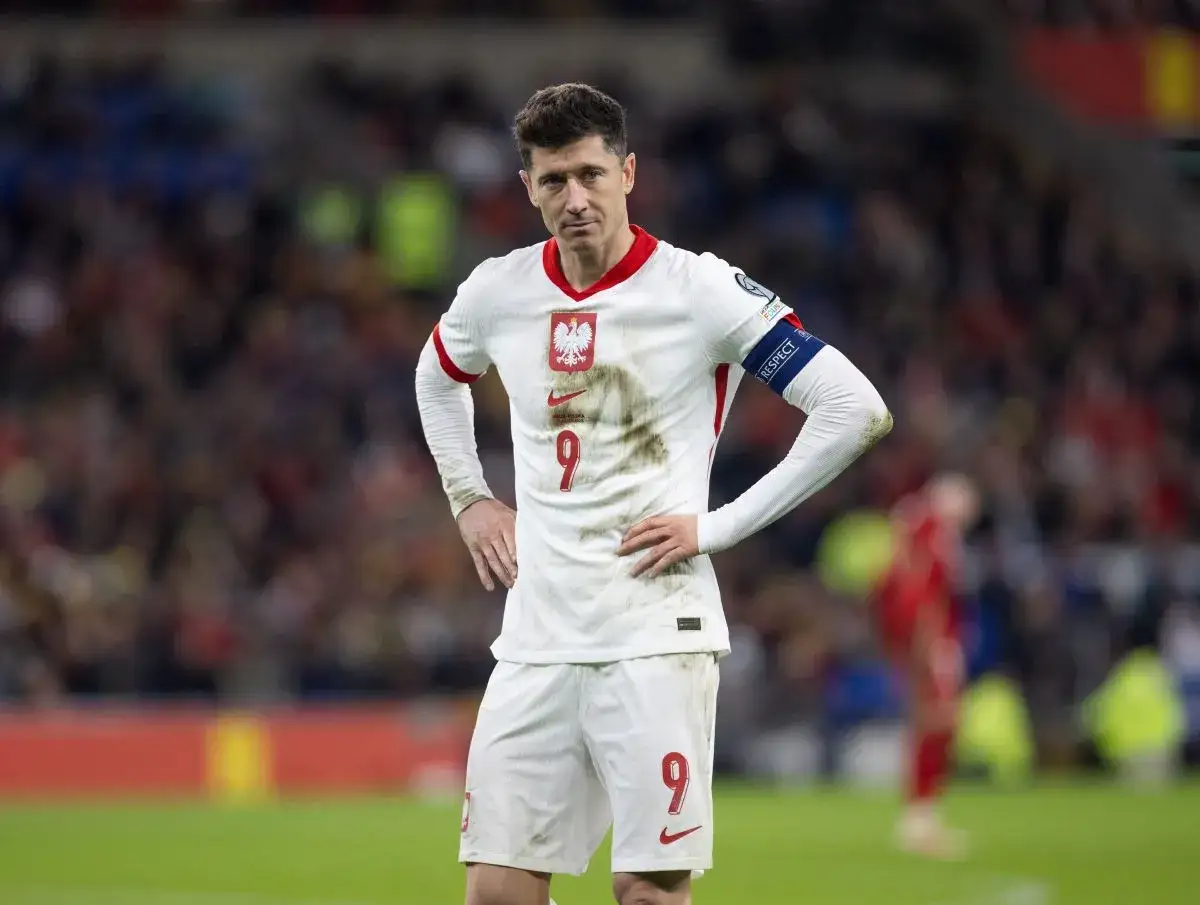 Robert Lewandowski w białej koszulce reprezentacji Polski, z numerem 9 na piersi i spodenkach, z rękami na biodrach.