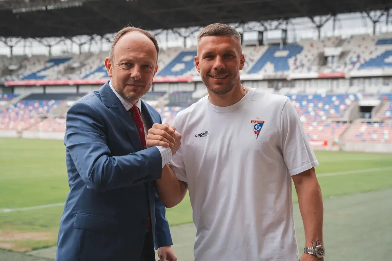 Dwóch mężczyzn ściska sobie dłonie na tle stadionu. Jeden w garniturze, drugi w koszulce z herbem Górnika Zabrze. Lukas Podolski, którego majątek budowany jest na sukcesach sportowych, pozuje do zdjęcia.
