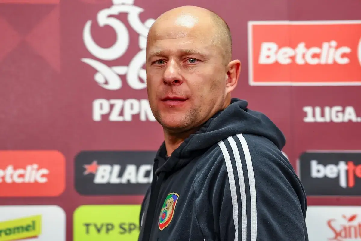 Łysy mężczyzna w bluzie z trzema paskami, z emblematem klubu na piersi. To trener Miedzi Legnica, gotowy do konferencji prasowej.