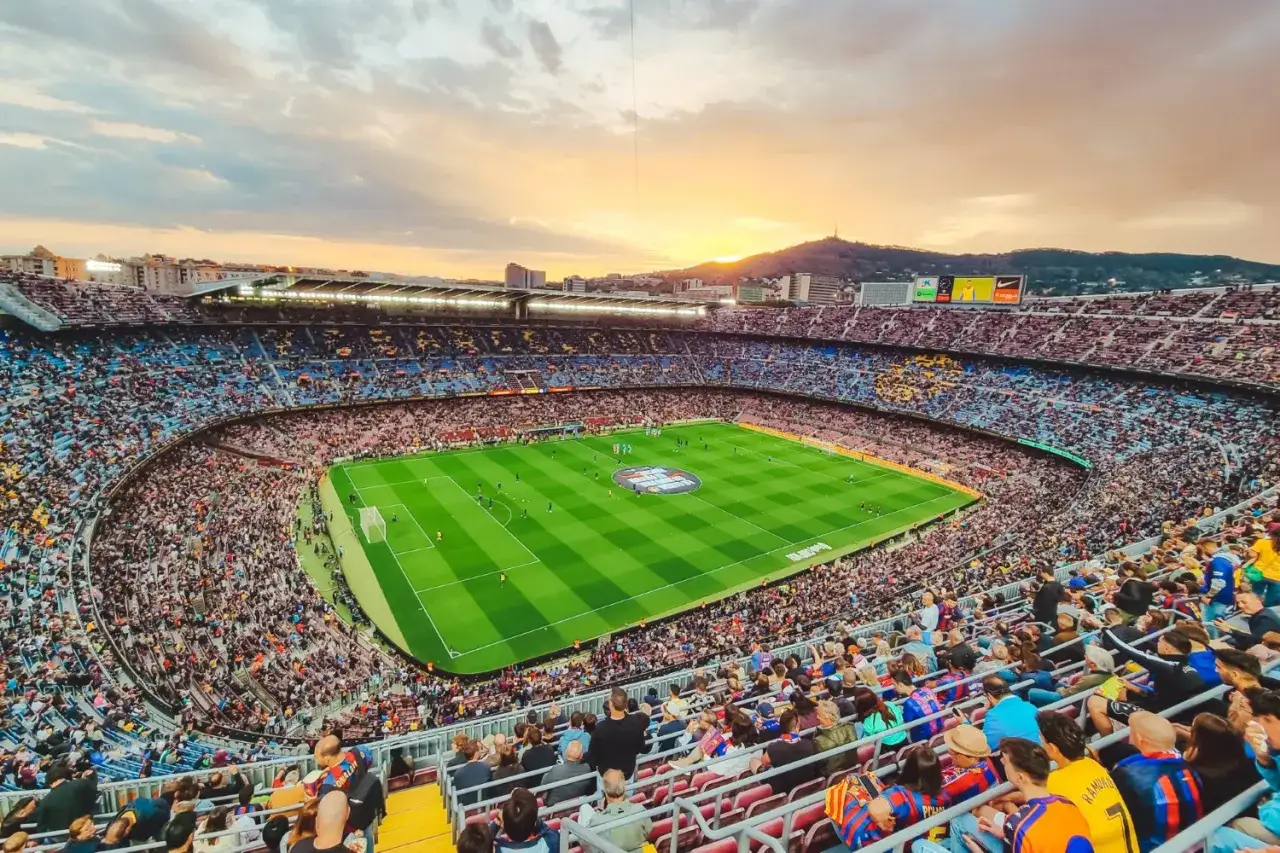 Zachód słońca nad Camp Nou w Bardelonie, pełnym kibiców. Piłkarze na zielonej murawie stadionu.