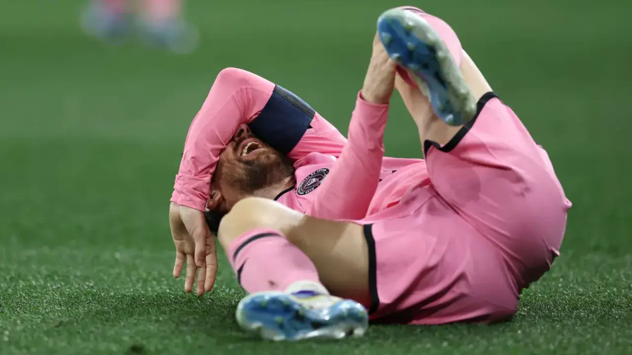Piłkarz w różowej koszulce leży na trawie, trzymając się za nogę. Wygląda na to, że Messi kontuzja uniemożliwiła mu dalszą grę.