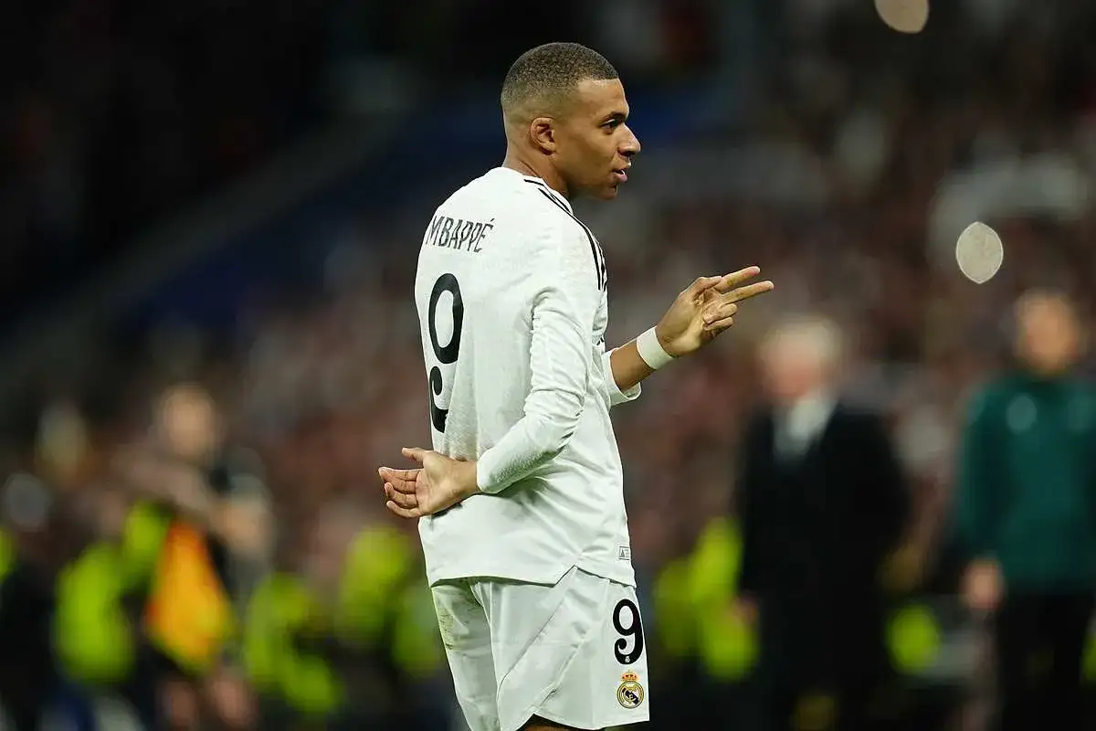 Mbappe, w białej koszulce z numerem 9, gestykuluje na boisku. Jego wzrost imponuje, a kibice śledzą każdy jego ruch.