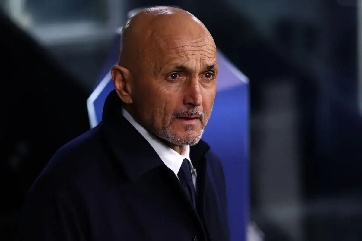 Łysy mężczyzna z brodą, ubrany w ciemny płaszcz i białą koszulę, wygląda na skupionego. To trener Juventusu, gotowy do poprowadzenia drużyny do zwycięstwa.