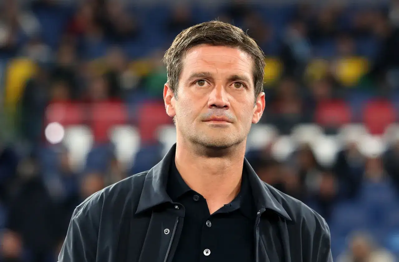 Mężczyzna w czarnej kurtce i koszulce polo, skupiony trener Interu, stoi na tle trybun stadionu.