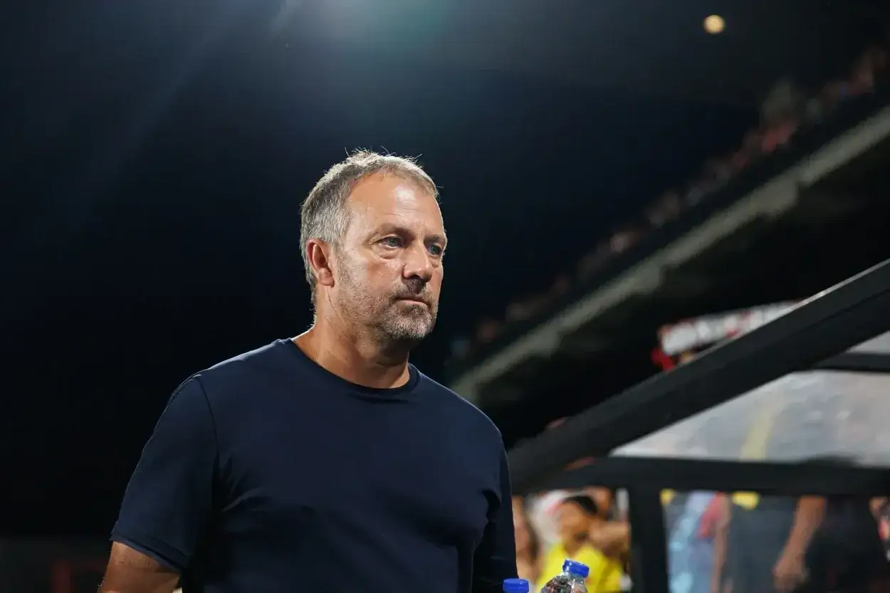 Hans-Dieter Flick, potencjalny trener Barcelony, wpatrzony w dal, z butelką wody w ręku, na tle nocnego stadionu.
