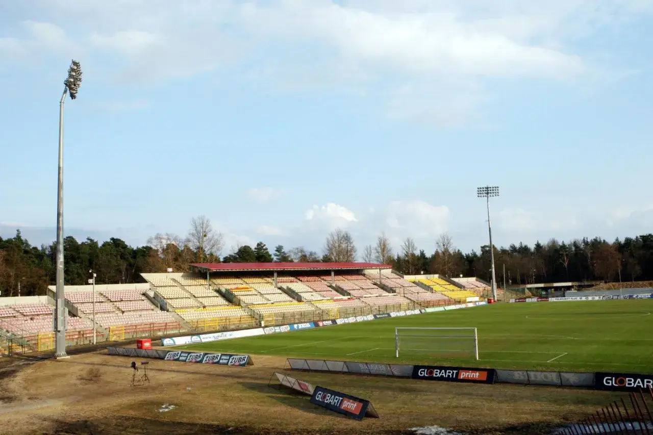 Puste trybuny starego stadionu Jagiellonii, z zieloną murawą i bramką. W tle las i niebo.