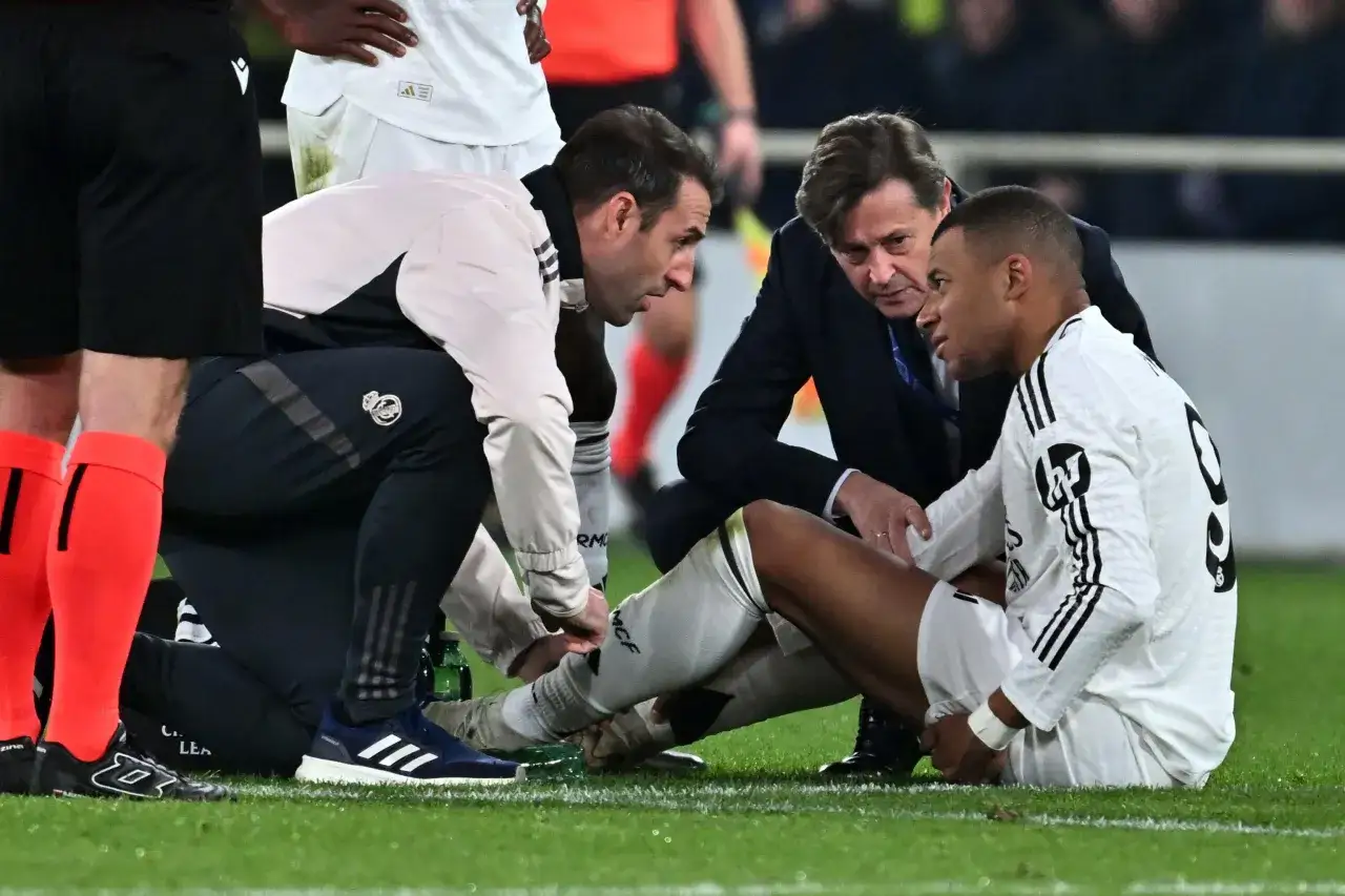 Mbappe kontuzja. Piłkarz siedzi na trawie, a sztab medyczny bada jego nogę.