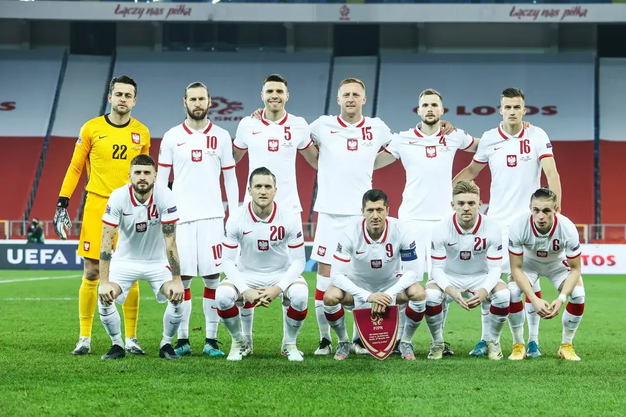 Piłkarze reprezentacji Polski w białych strojach pozują do zdjęcia na murawie. W pierwszej linii Robert Lewandowski z tarczą PZPN.