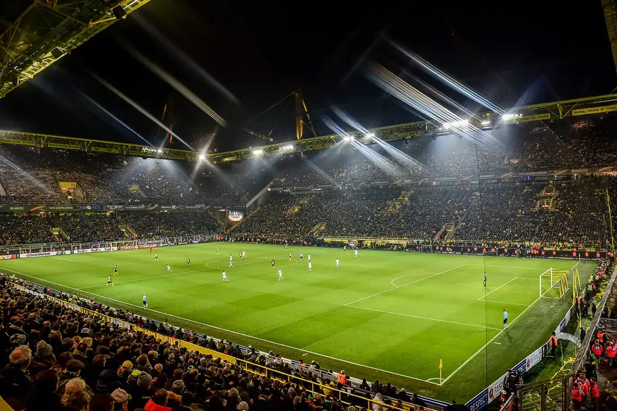 Nocny mecz na stadionie w Dortmundzie. Tysiące kibiców w żółtych barwach wypełnia trybuny, a piłkarze rozgrywają spotkanie przy sztucznym oświetleniu.