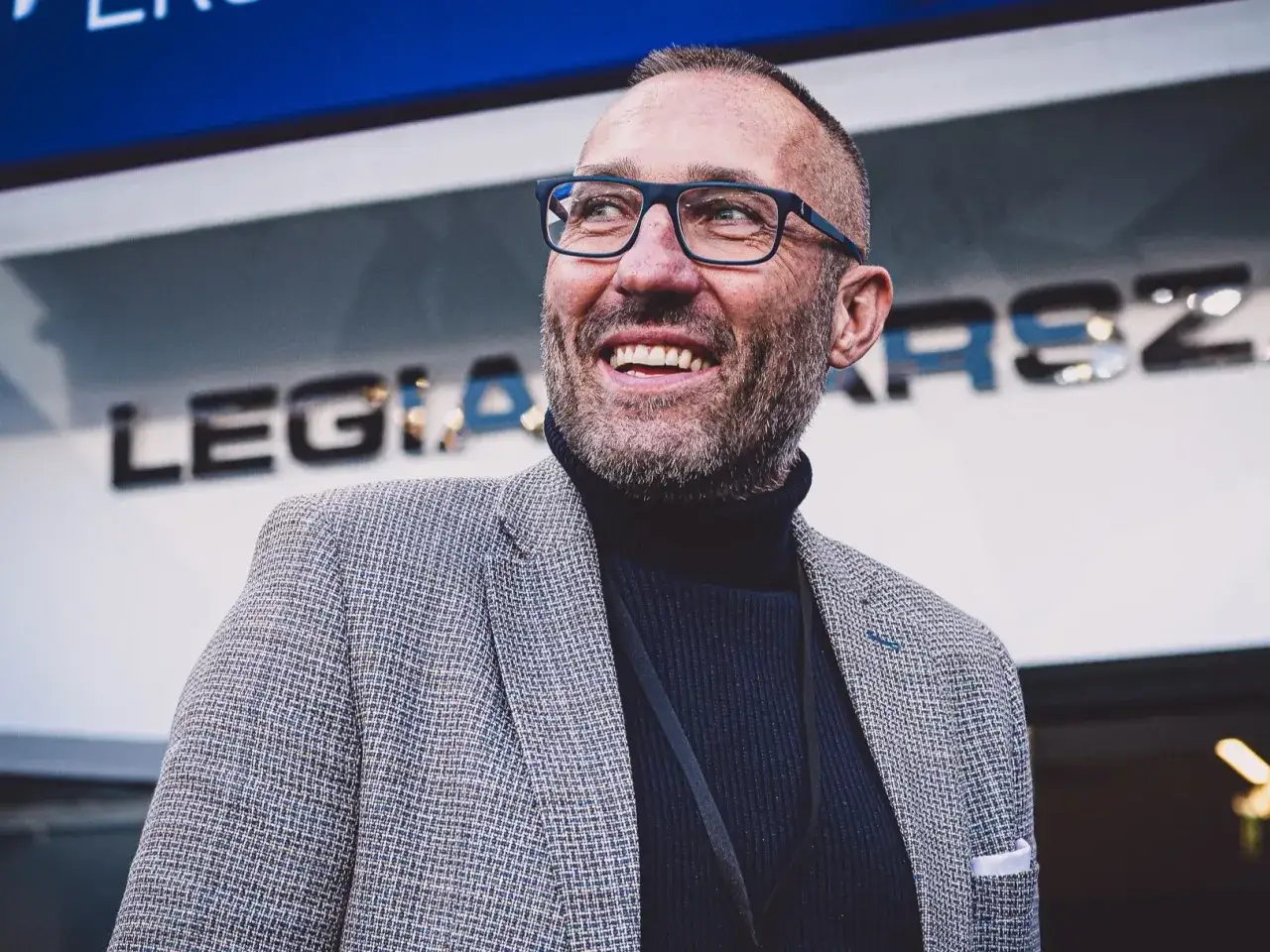 Uśmiechnięty Marek Papszun w okularach i marynarce, z logo Legii Warszawa w tle.