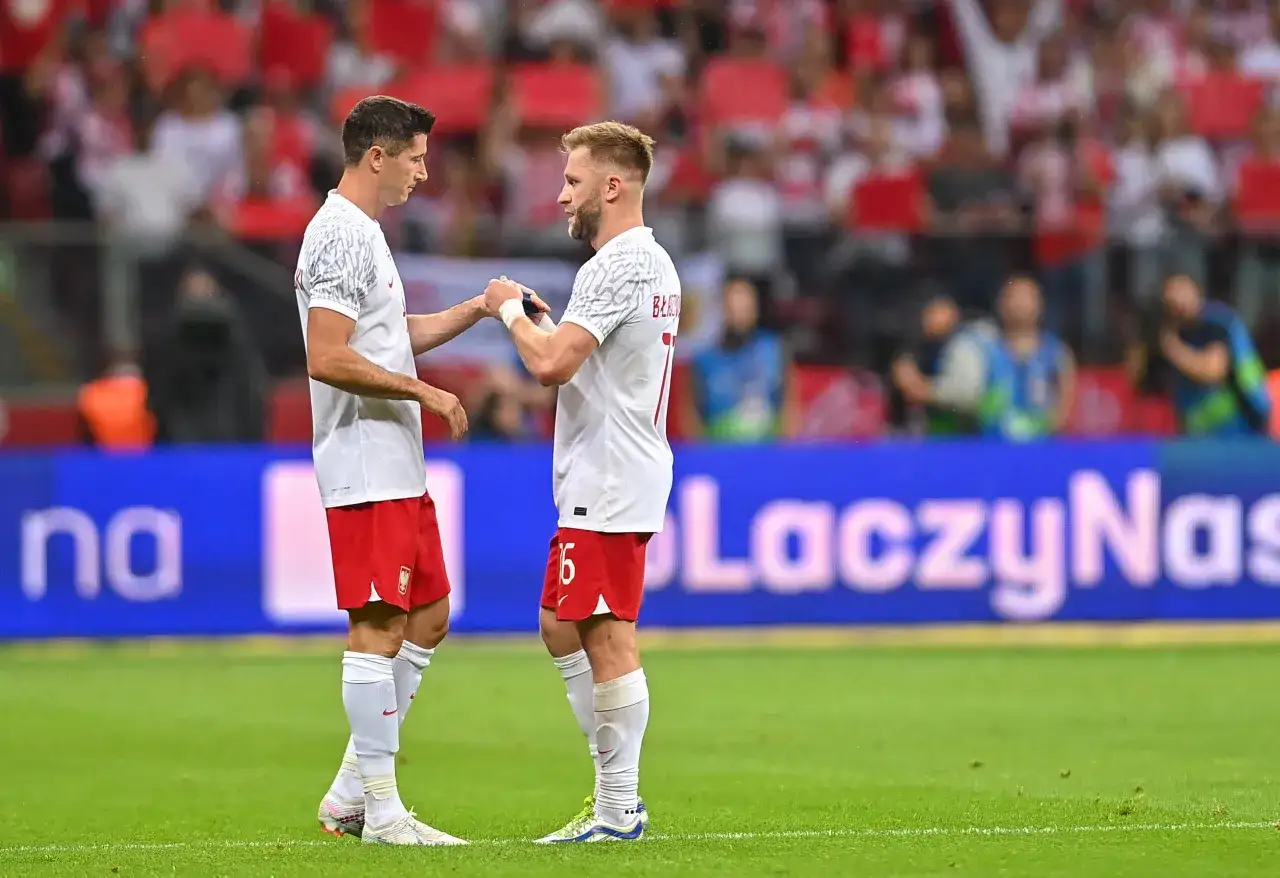 Piłkarze w białych strojach, Lewandowski i Błaszczykowski, wymieniają uścisk dłoni na boisku. Ich błyszczący wzrost inspiruje kibiców.