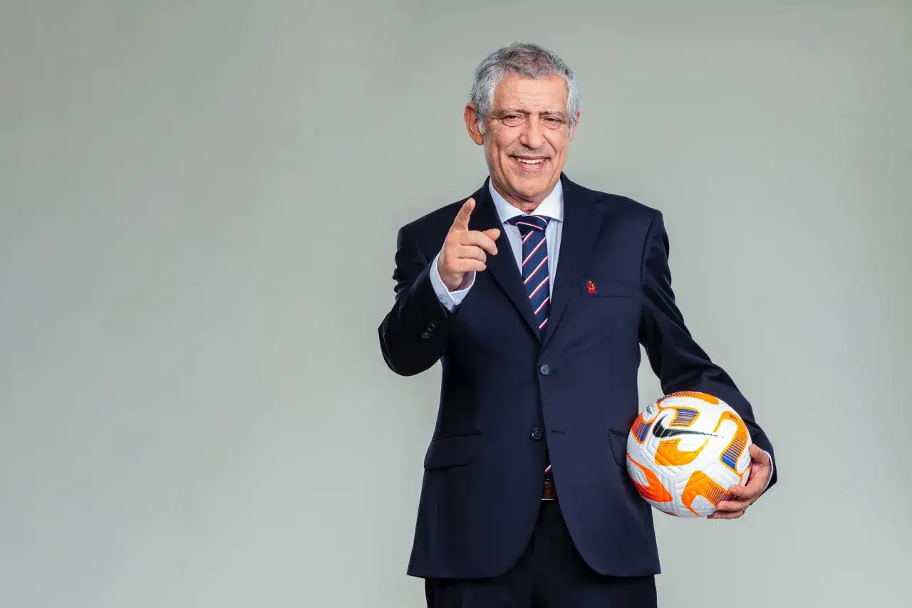Uśmiechnięty mężczyzna w garniturze trzyma piłkę, wskazując palcem. To inspiracja, co można trenować na kursie UEFA B.