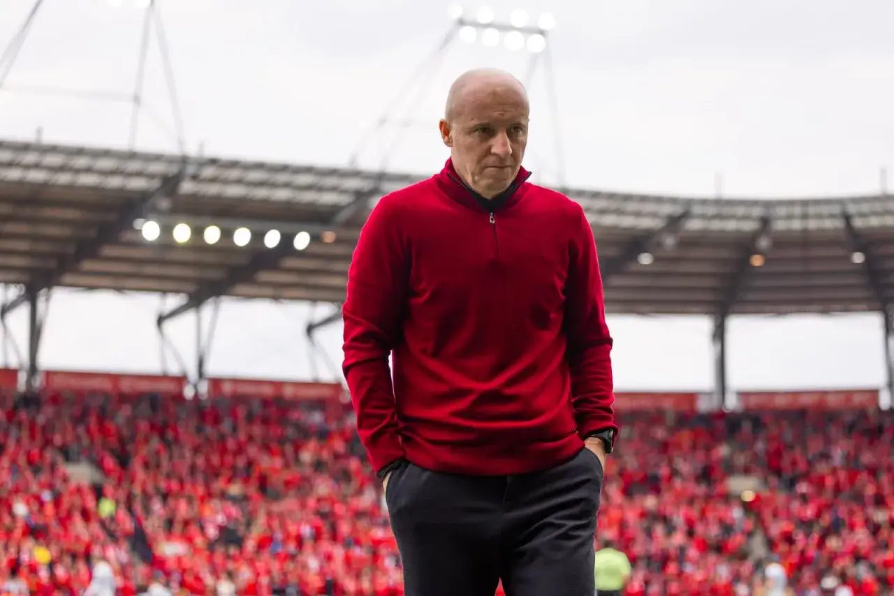 Nowy trener Widzewa, w czerwonym swetrze, idzie po murawie stadionu, na którym zasiadają kibice w czerwonych barwach.