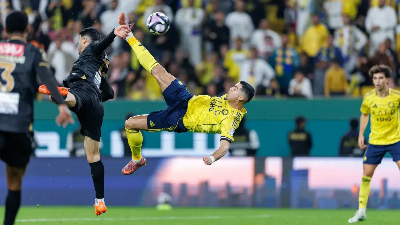 Piłkarz w żółtej koszulce wykonuje przewrotkę, próbując trafić w piłkę. Gra w klubie Al Nassr, gdzie gra Ronaldo.