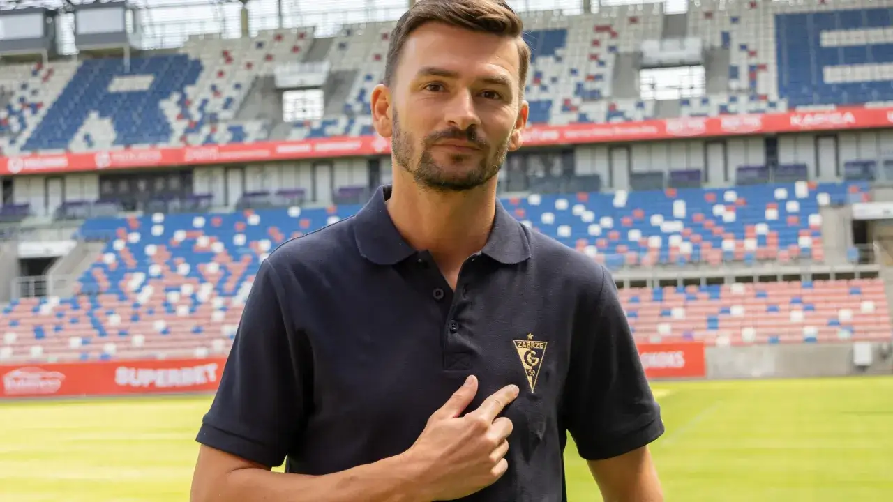 Trener Górnika Zabrze, z dumą prezentuje herb klubu na koszulce. Stadion w tle.