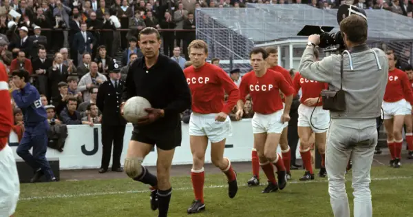 Bramkarz w czarnym stroju trzyma piłkę, obok niego biegną zawodnicy w czerwonych koszulkach z napisem CCCP. Kamera filmowa rejestruje mecz.